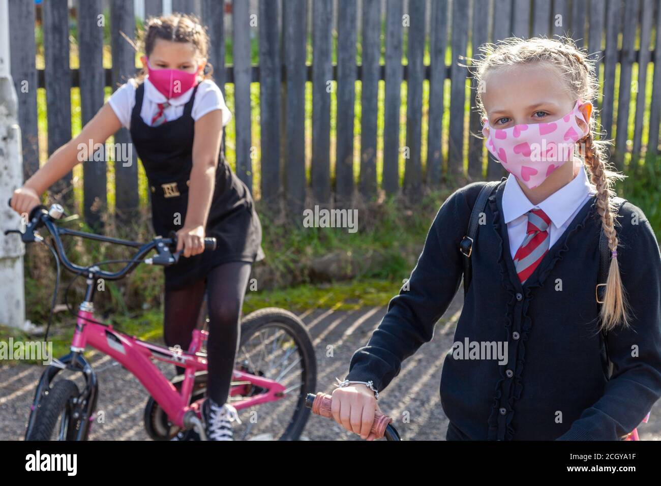 Two ten year old school friends in school uniforms sitting on bicycles and wearing face masks. Stock Photo