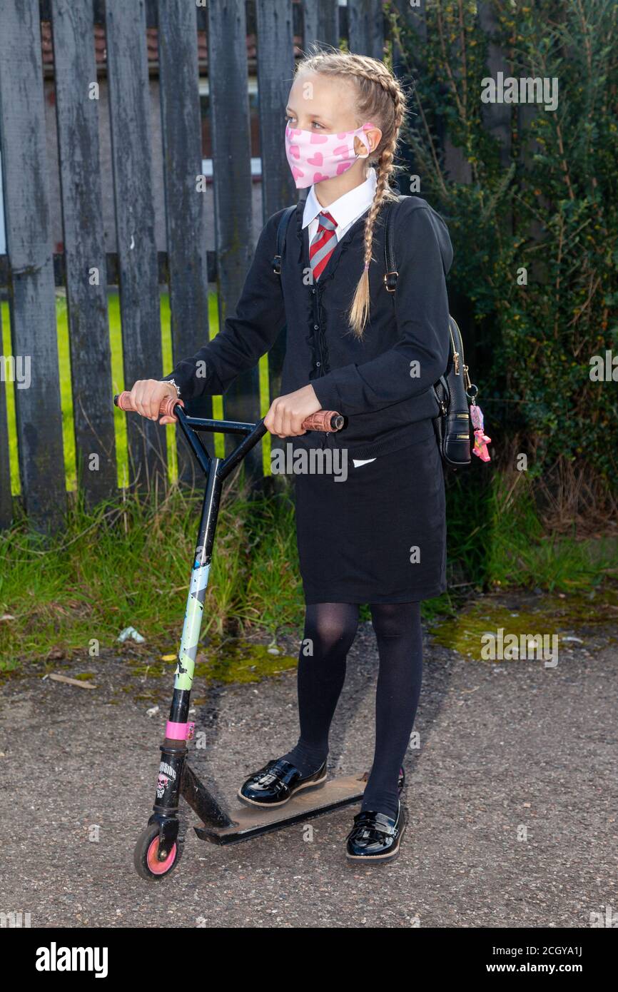 A 10 year old schoolgirl in school uniform wearing a face mask standing with one foot on her scooter Stock Photo