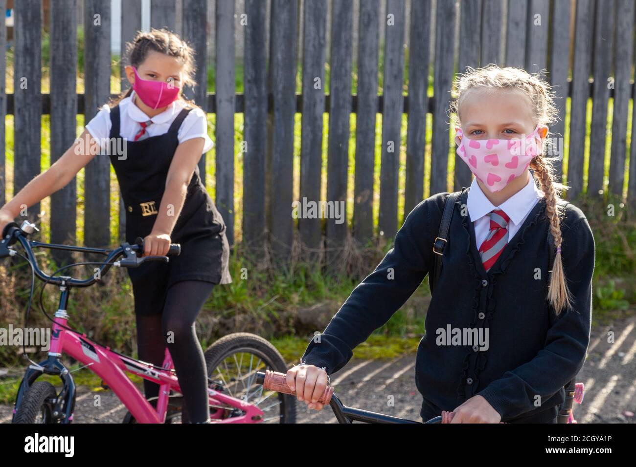 Two ten year old school friends in school uniforms sitting on bicycles and wearing face masks. Stock Photo