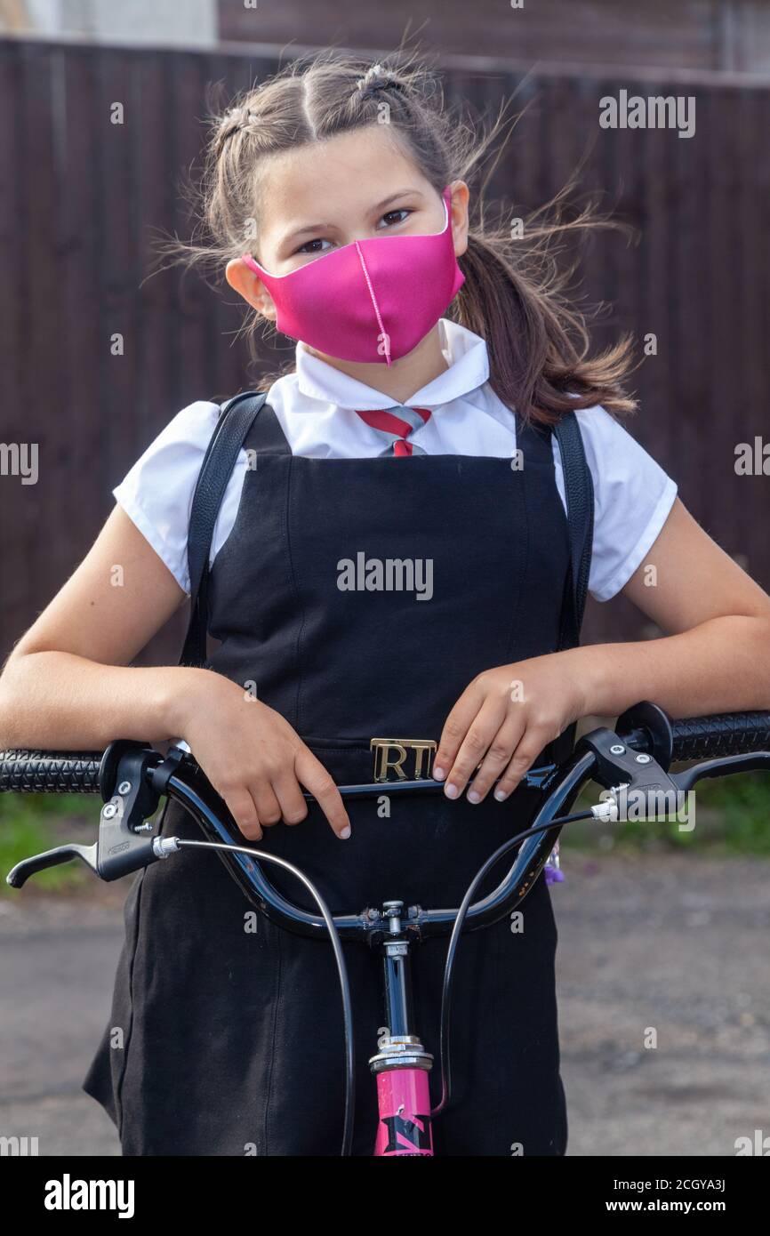 A 10 year old schoolgirl in school uniform and wearing a face mask standing with her elbows on the handlebars of her pink bike. Stock Photo