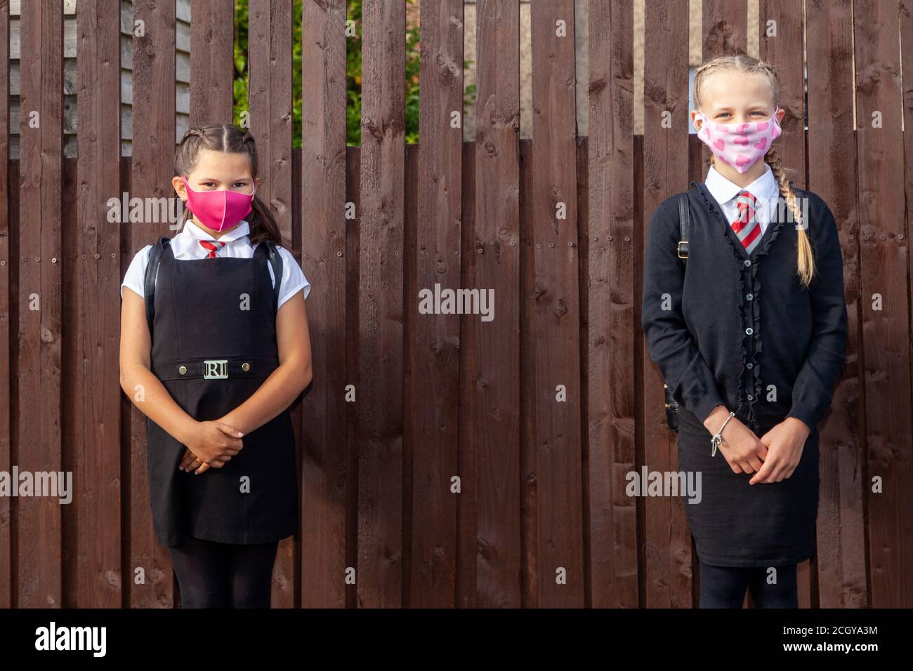 Two 10 year old schoolgirls wearing school uniforms and face masks and standing next to a wooden fence Stock Photo