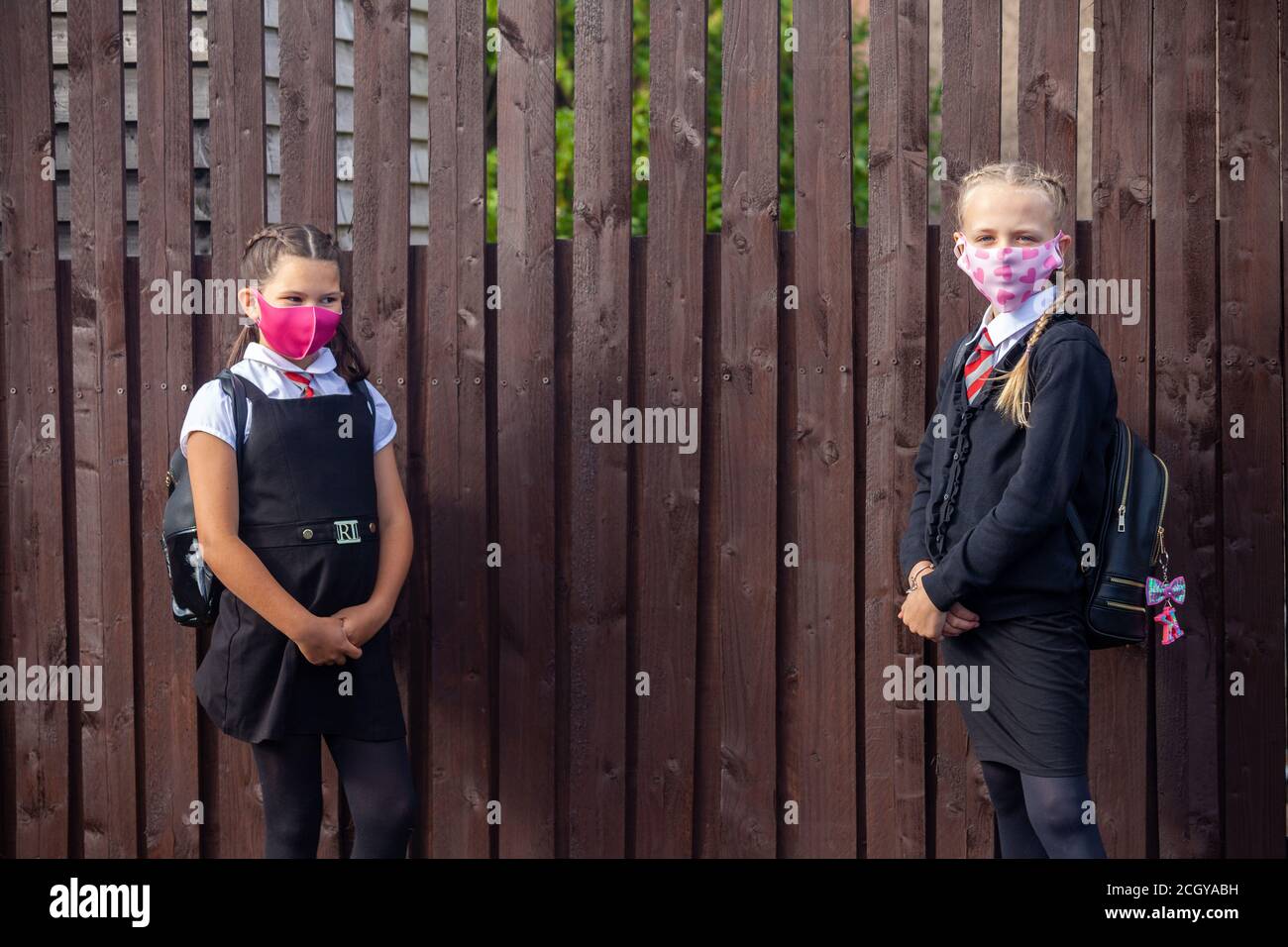 Two 10 year old schoolgirls wearing school uniforms and face masks and standing next to a wooden fence Stock Photo