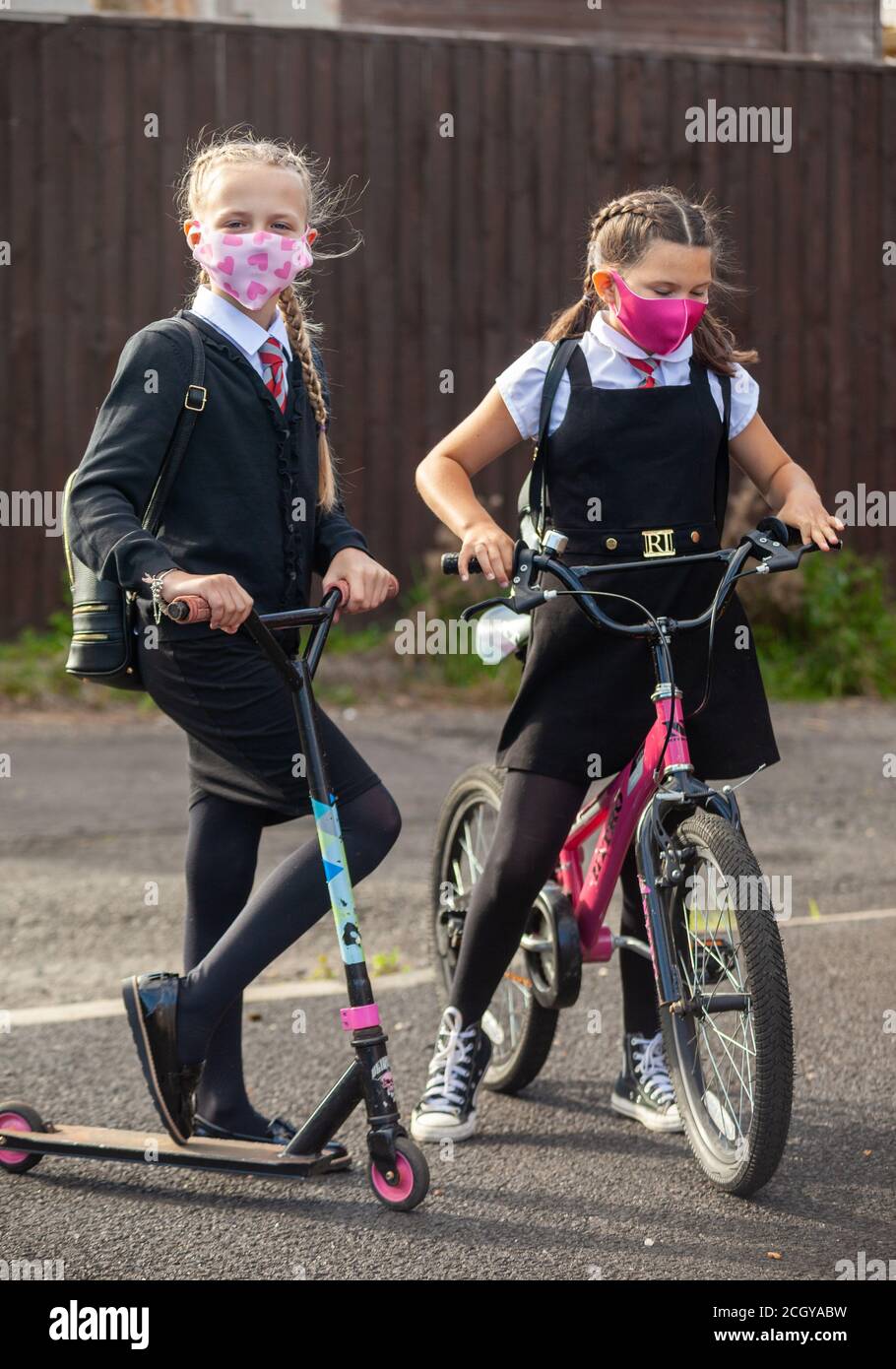 Two schoolgirls in school uniform standing with their bike and scooter and wearing face masks. Stock Photo