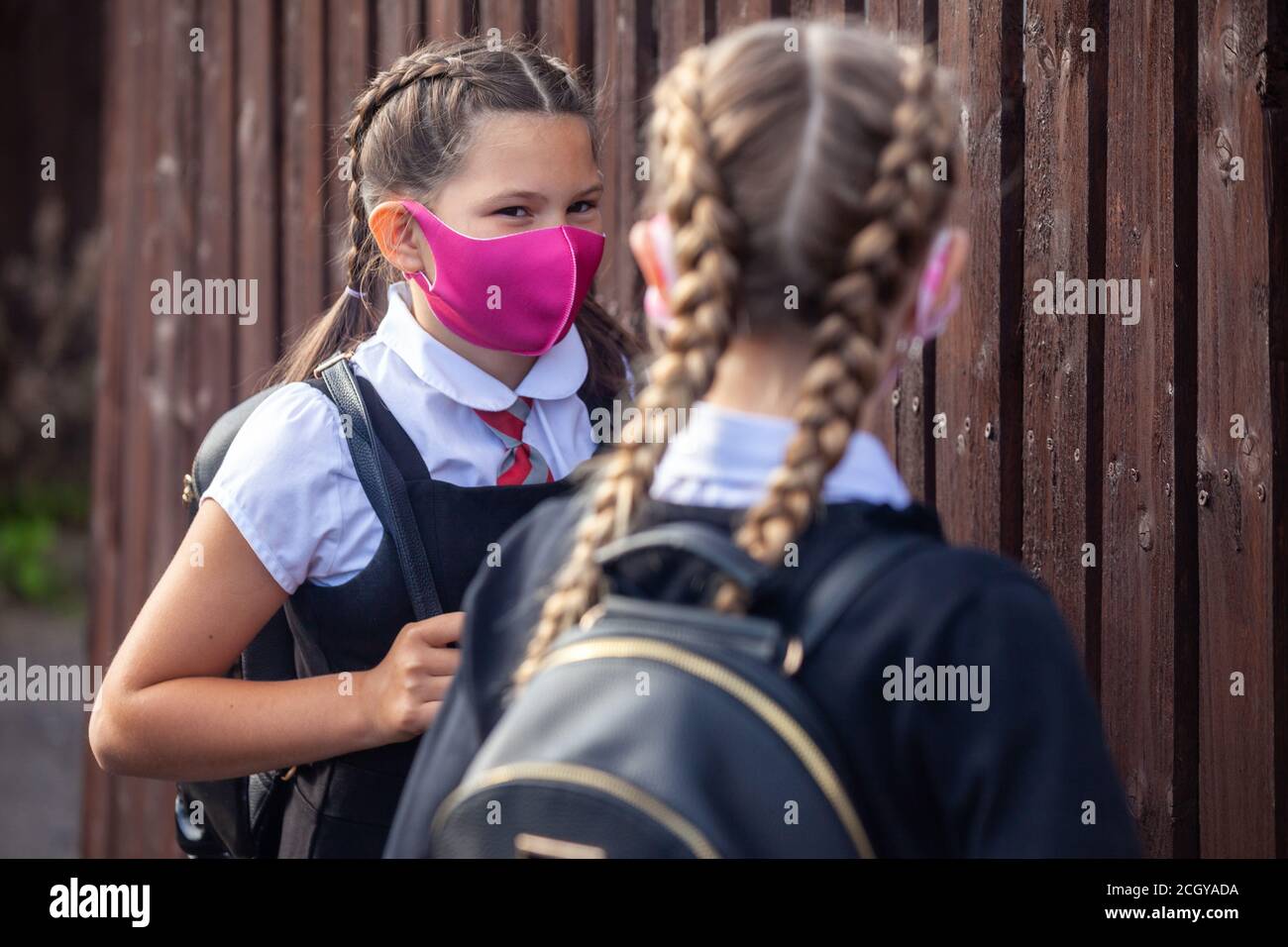 Two 10 year old school friends chatting to each other next to a wooden fence and wearing face masks. Stock Photo