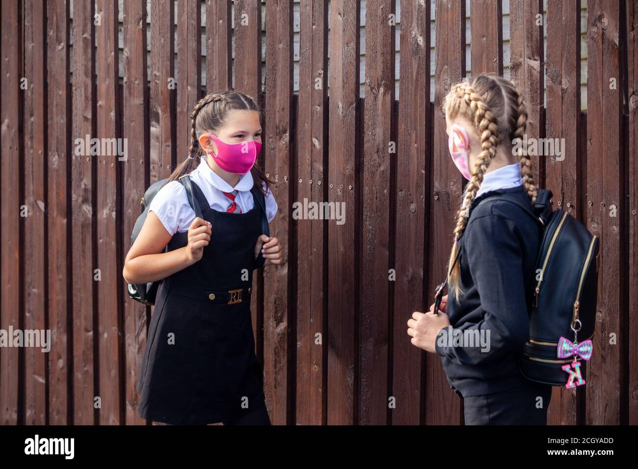 Two 10 year old school friends chatting to each other next to a wooden fence and wearing face masks. Stock Photo