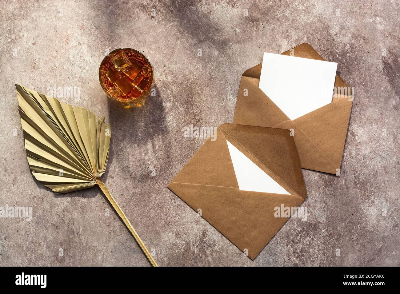 Whiskey with ice in a glass, blank paper mockup cards in a craft envelope, dry palm leaf fan in the sunlight. Male background. Alcohol industry templa Stock Photo