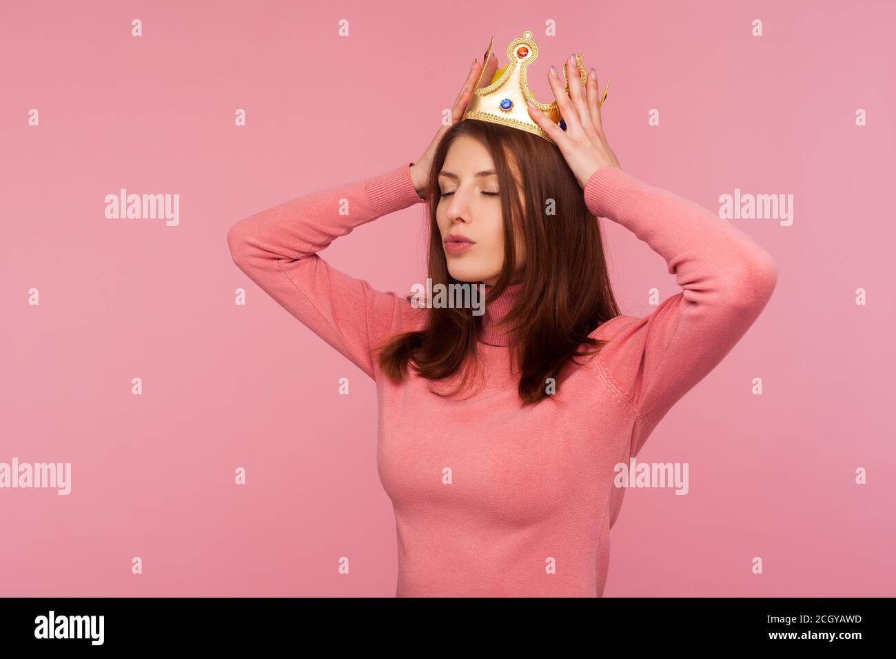 Selfish confident brunette woman adjusting golden crown on her head, proud of her status, pretending to be queen. Indoor studio shot isolated on pink Stock Photo