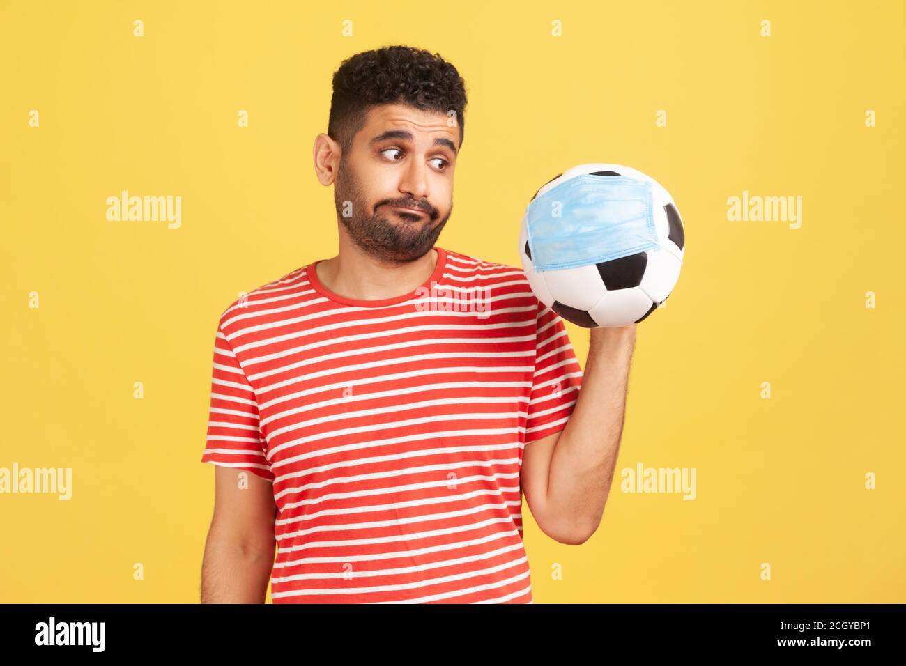 Upset man in red t-shirt looking at soccer ball in safety face mask with disappointment, nervous because of football championship results falsificatio Stock Photo