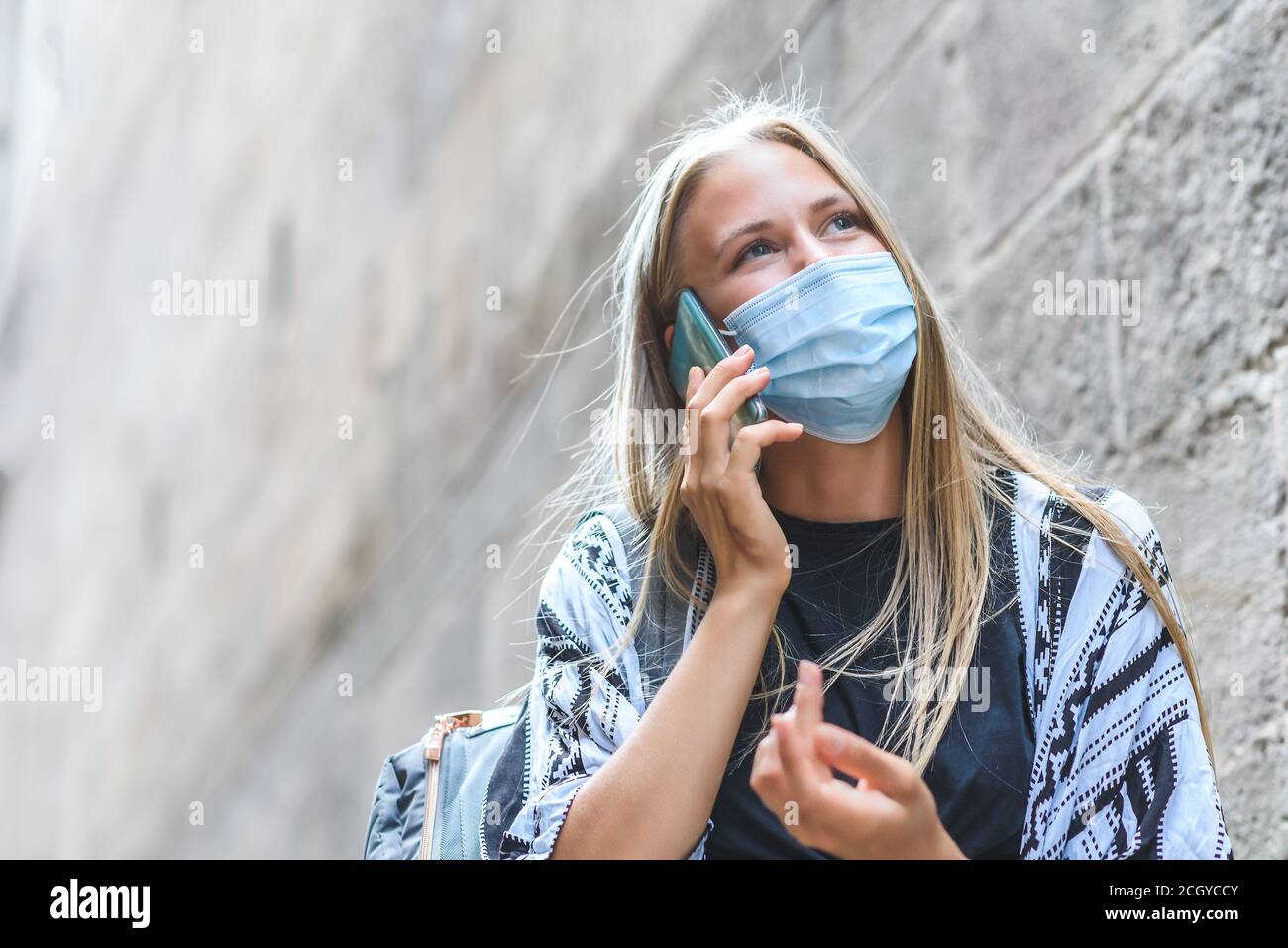 Portrait of a beautiful blonde caucasian woman using a blue face mask talking by phone near an ancient building. Copy Space on the left Stock Photo