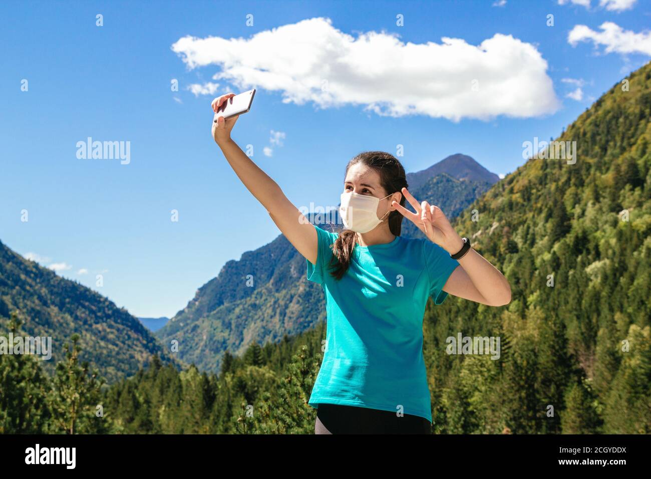 Stock photo of a 30's woman with face mask taking a selfie with phone while enjoying the mountains and a beautiful landscapes on a sunny day Stock Photo