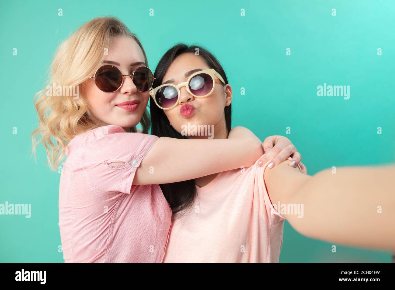 Portrait of pretty charming girls in casual outfits, wearing sunglasses shooting selfie , making duck faces, using smart phone isolated on studio blue Stock Photo