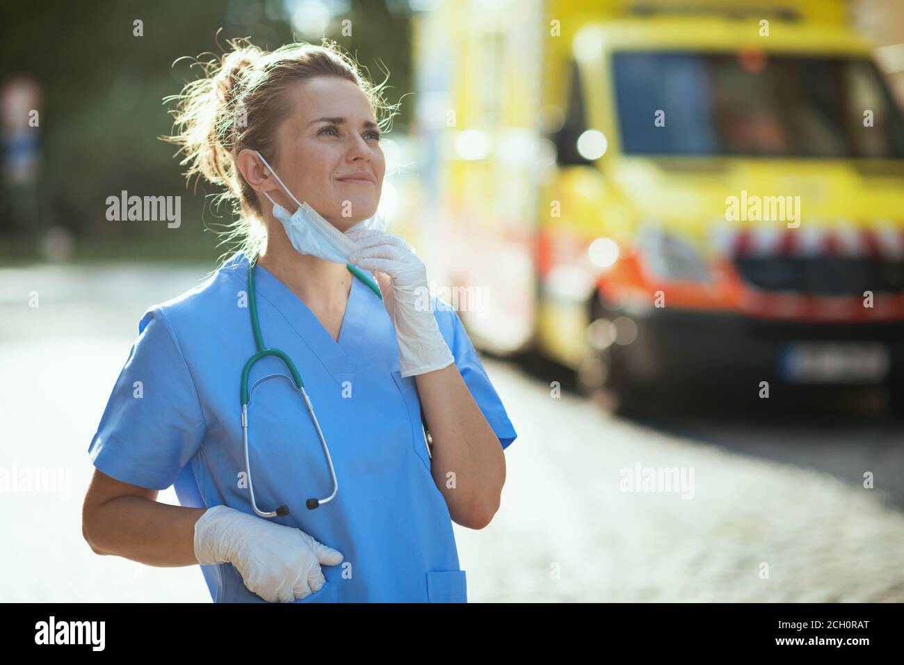 covid-19 pandemic. relaxed modern medical doctor woman in scrubs with stethoscope and medical mask breathing outdoors near ambulance. Stock Photo