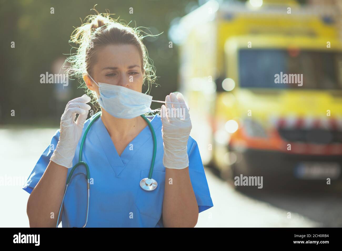 covid-19 pandemic. tired modern medical doctor woman in scrubs with stethoscope and medical mask outside near ambulance. Stock Photo