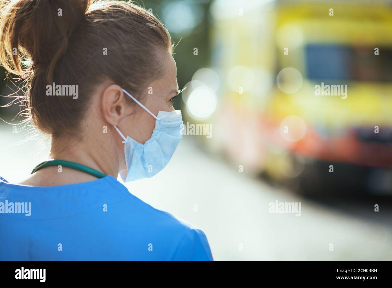 covid-19 pandemic. Seen from behind medical doctor woman in uniform with medical mask outside near ambulance. Stock Photo