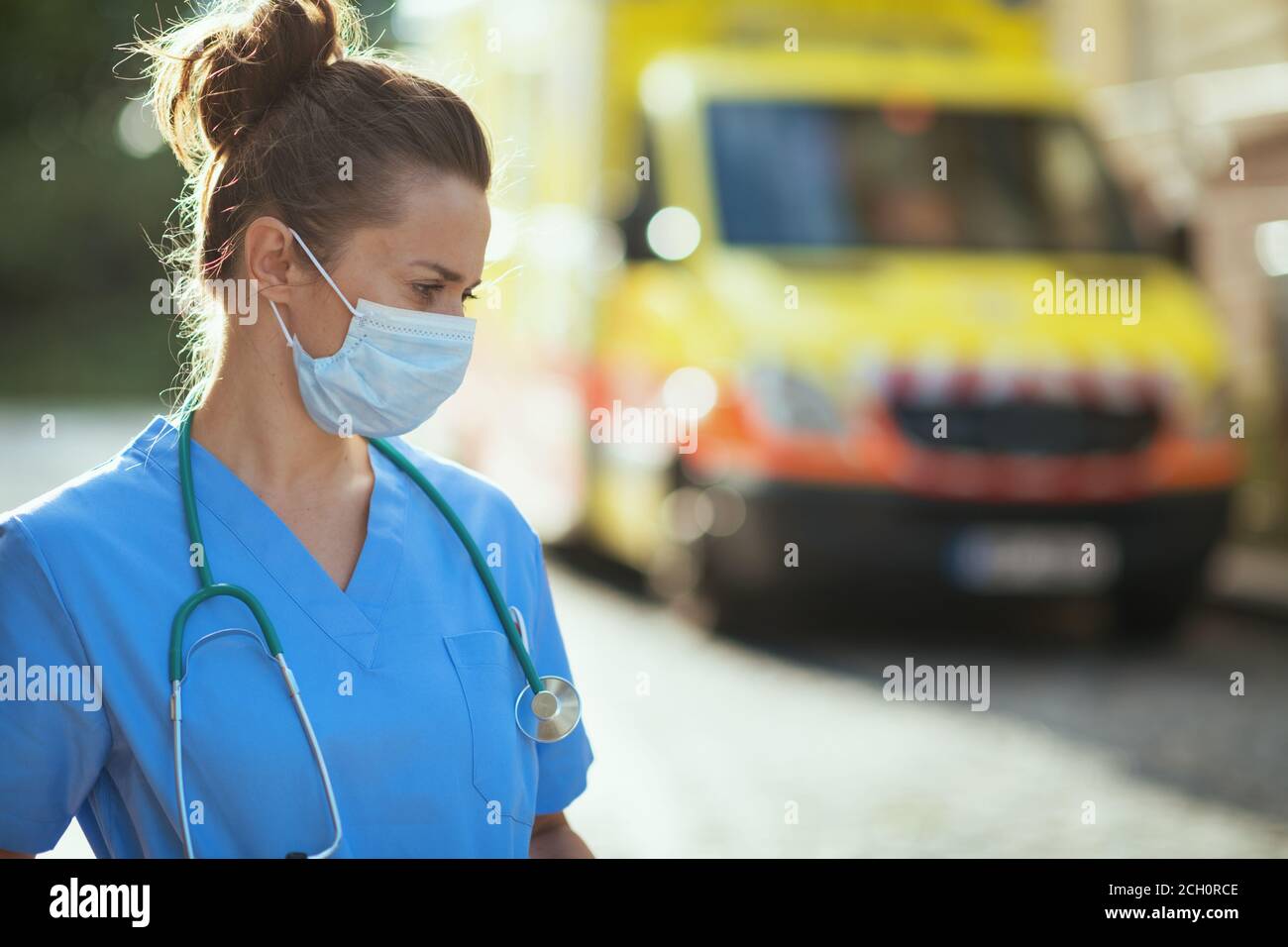 coronavirus pandemic. pensive modern paramedic woman in scrubs with stethoscope and medical mask outside near ambulance. Stock Photo