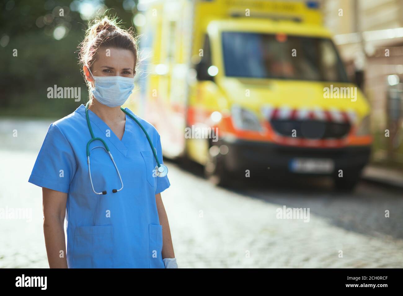 covid-19 pandemic. Portrait of modern paramedic woman in uniform with stethoscope and medical mask outside near ambulance. Stock Photo