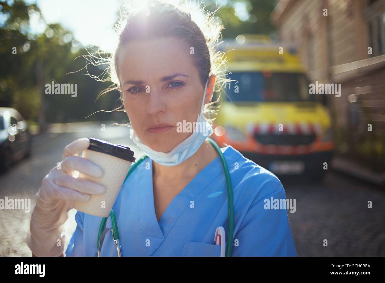 covid-19 pandemic. tired modern paramedic woman in uniform with stethoscope, medical mask and cup of coffee outside near ambulance. Stock Photo