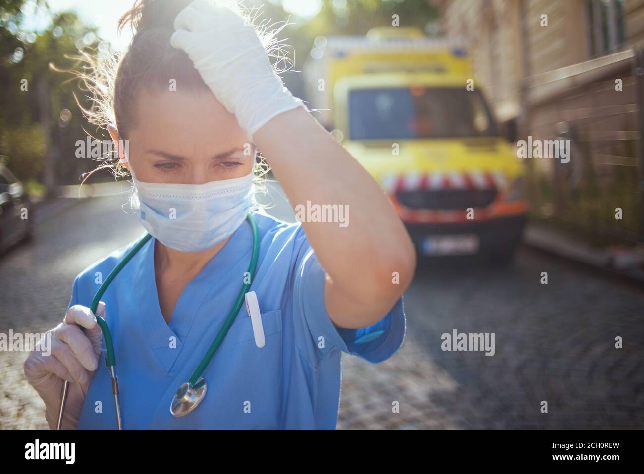 coronavirus pandemic. sad modern medical doctor woman in scrubs with stethoscope and medical mask outdoors near ambulance. Stock Photo