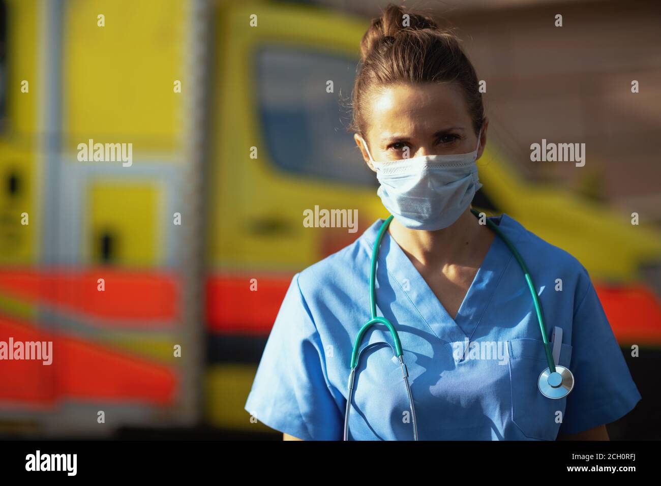 covid-19 pandemic. Portrait of modern medical doctor woman in scrubs with stethoscope and medical mask outside near ambulance. Stock Photo