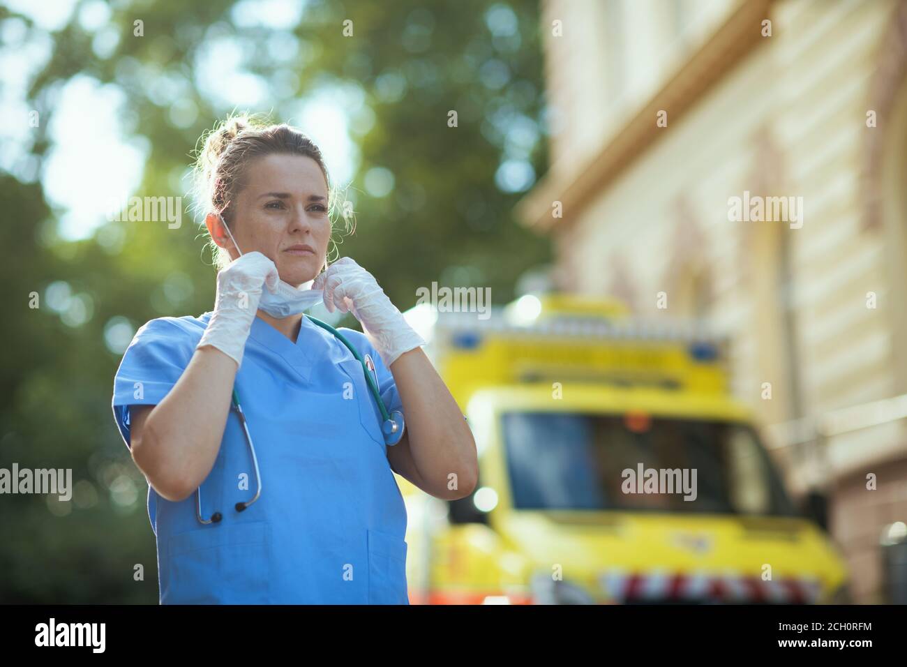 covid-19 pandemic. modern paramedic woman in scrubs with stethoscope and medical mask outdoors near ambulance. Stock Photo