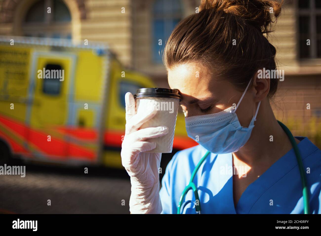 coronavirus pandemic. tired modern paramedic woman in uniform with stethoscope, medical mask and cup of coffee outside near ambulance. Stock Photo