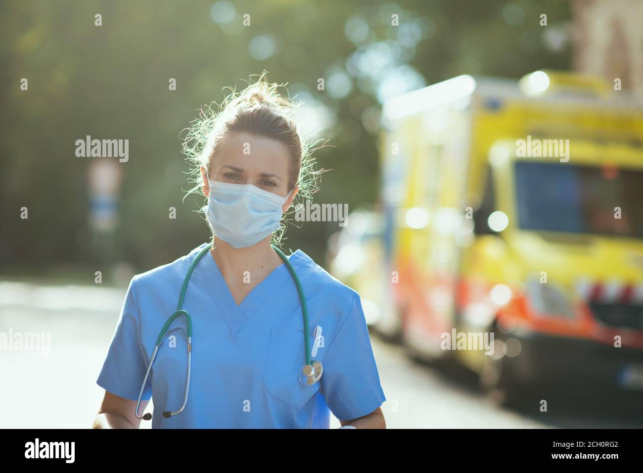 covid-19 pandemic. Portrait of modern paramedic woman in scrubs with stethoscope and medical mask outdoors near ambulance. Stock Photo