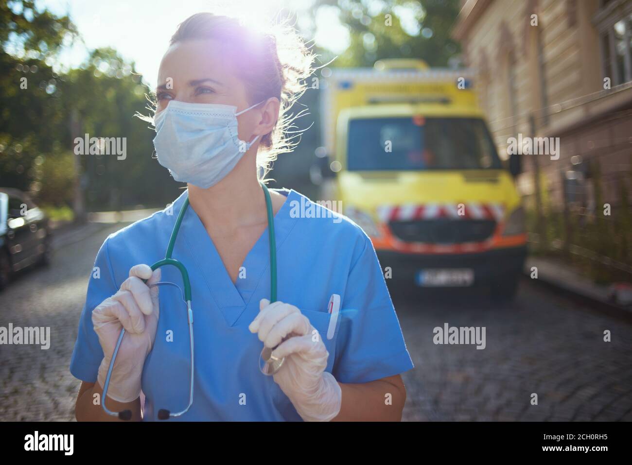 coronavirus pandemic. modern paramedic woman in uniform with stethoscope and medical mask looking into the distance outside near ambulance. Stock Photo