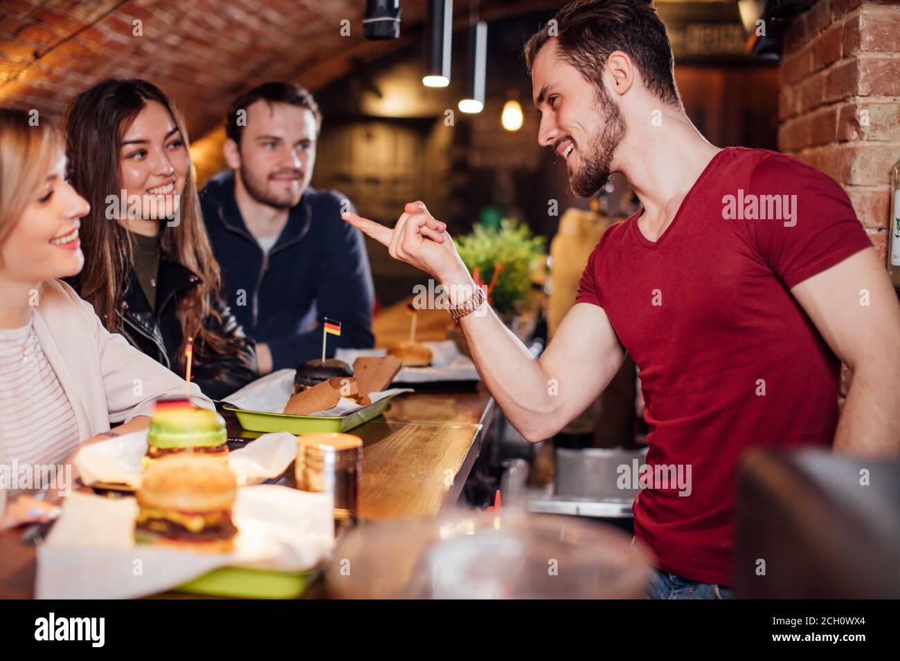 Young attractive barkeeper flirting and joking with female visitors sitting at bar counter and having lunch with smiling male friends Stock Photo