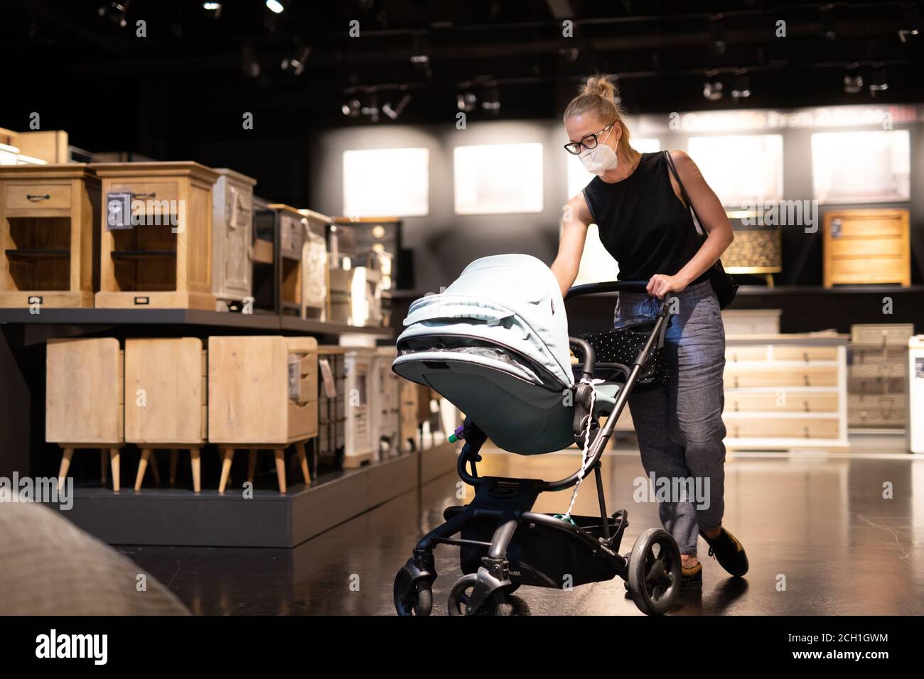Young mom with newborn in stroller shopping at retail furniture and home accessories store wearing protective medical face mask to prevent spreading Stock Photo