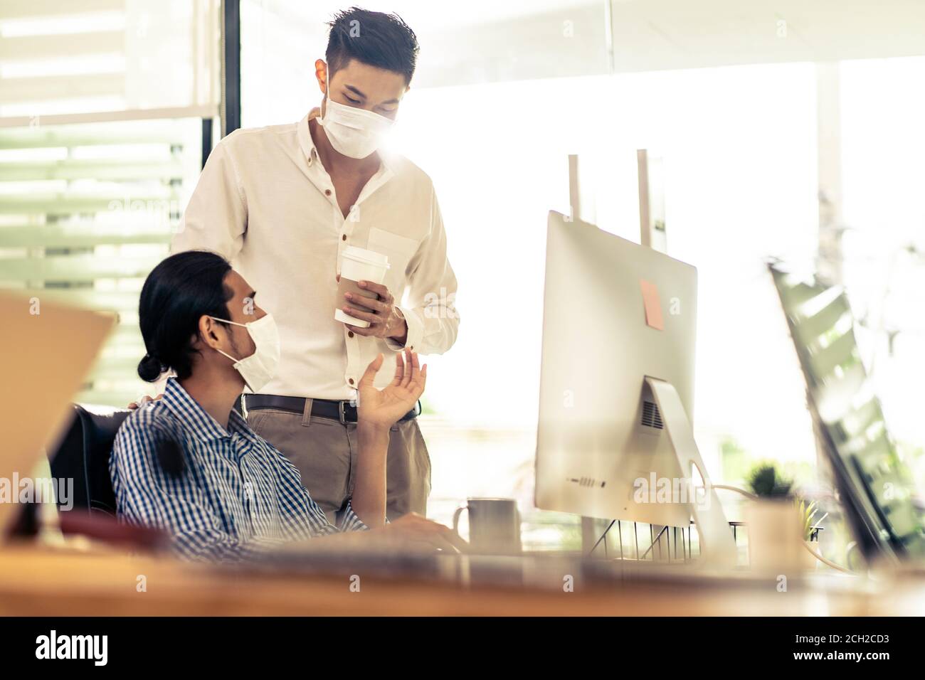 Two asian businessman talk discuss their work in morning after office reopen due to coronavirus COVID-19 pandemic. They wear protective face mask to p Stock Photo