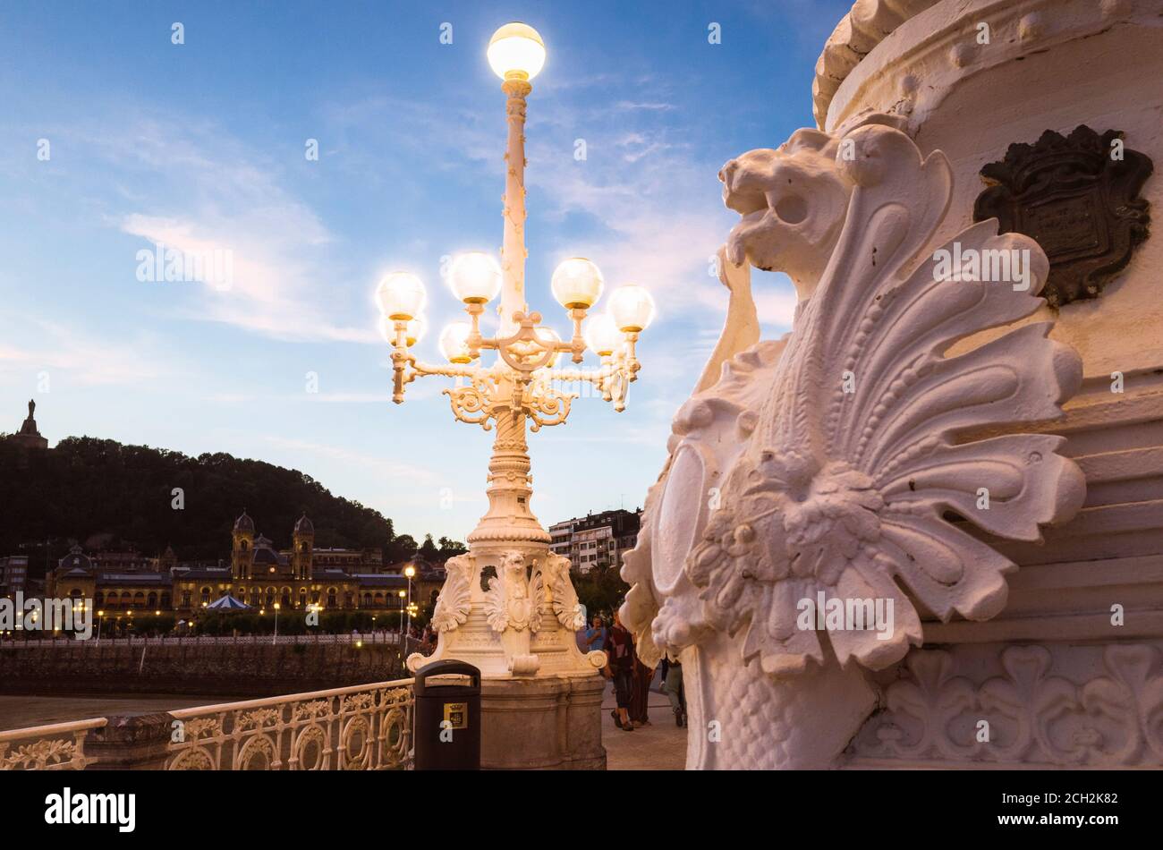 Donostia, Gipuzkoa, Basque Country, Spain - July 12th, 2019 : Close up of ornated street lamps at dusk with incidental people and city hall in backgro Stock Photo