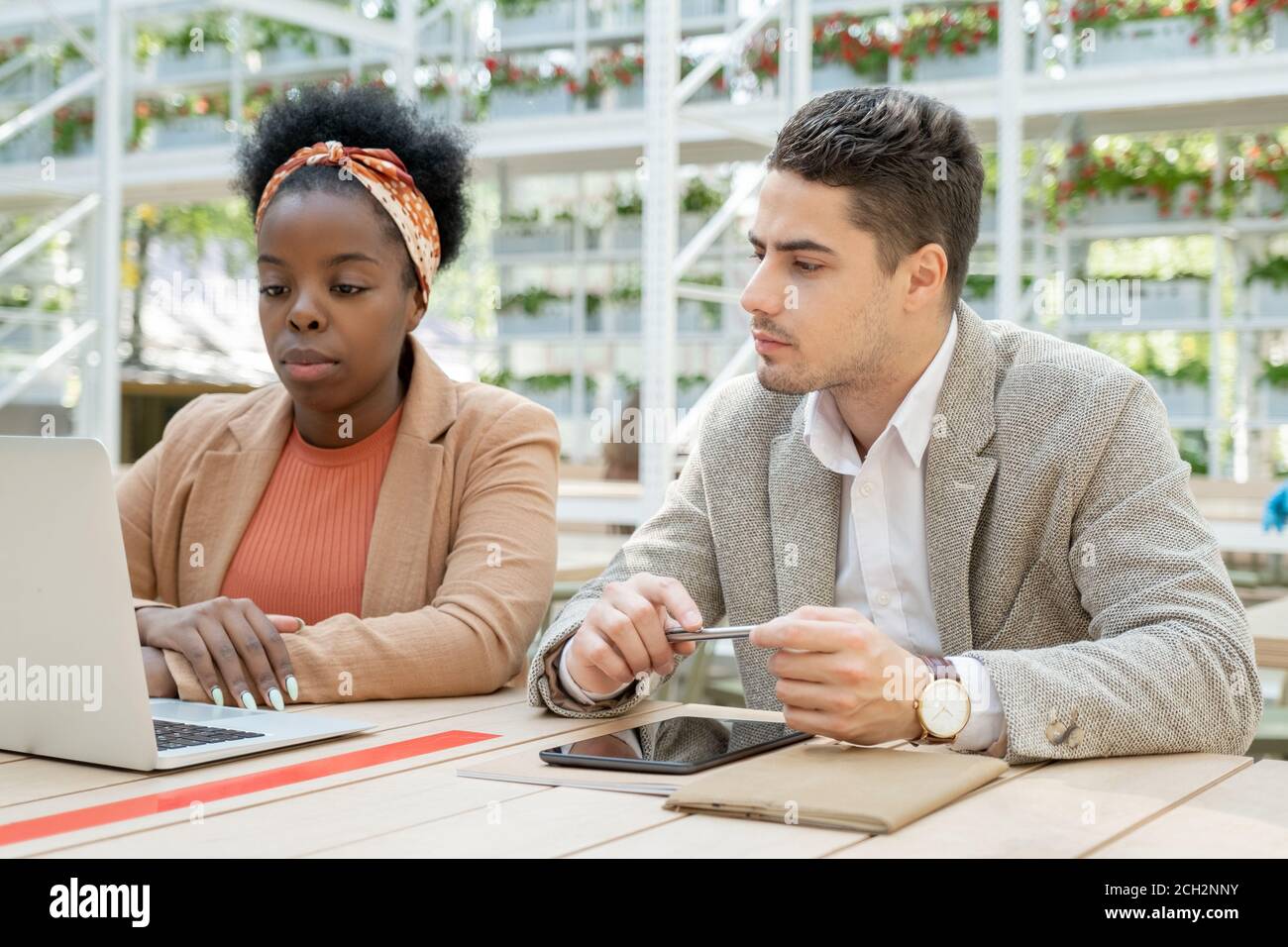 Two young serious intercultural colleagues watching online training by table Stock Photo