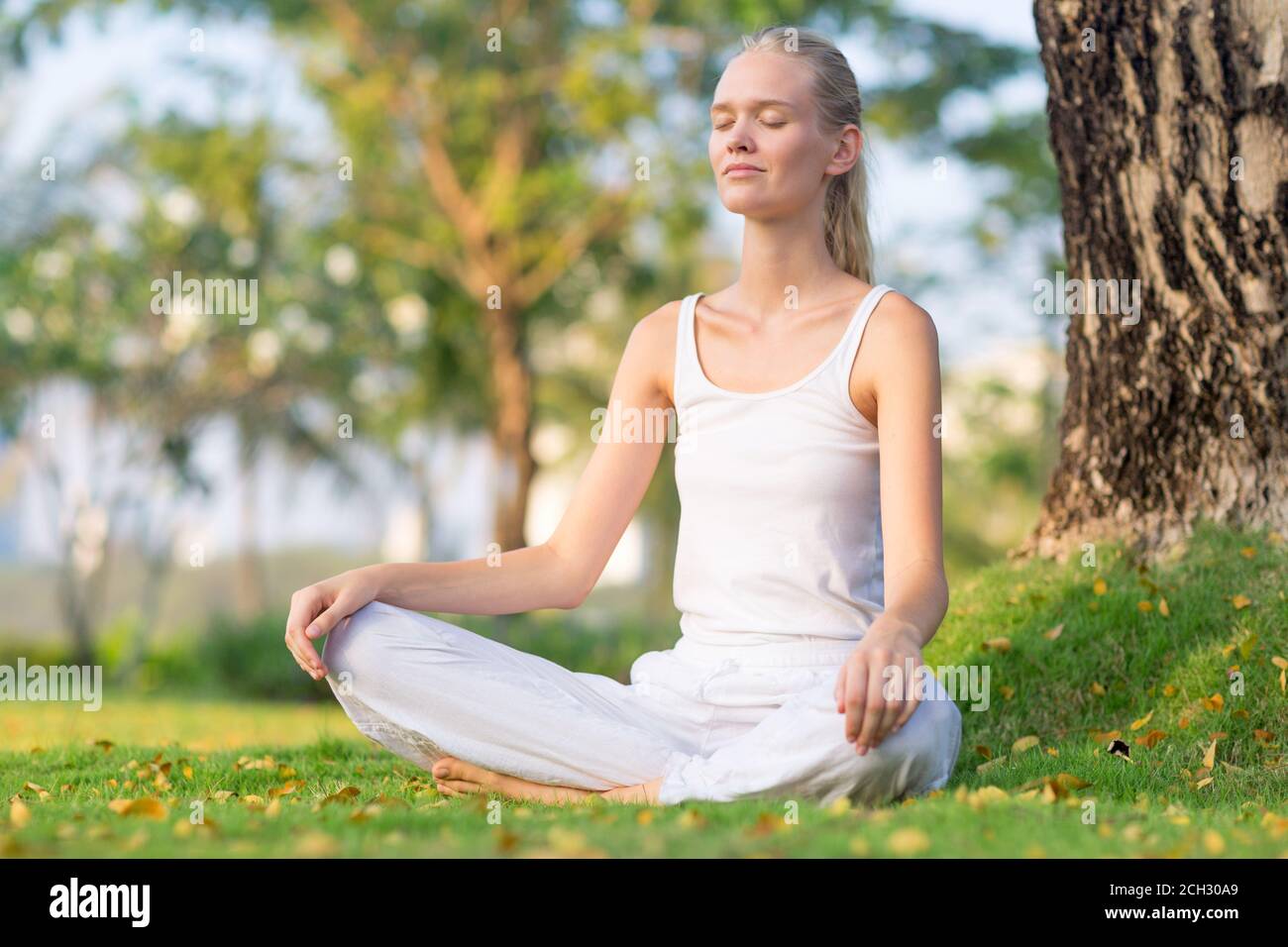 Relaxed young woman meditating at peace outside sitting at the park during summer. Wellness, health and self care. Stock Photo