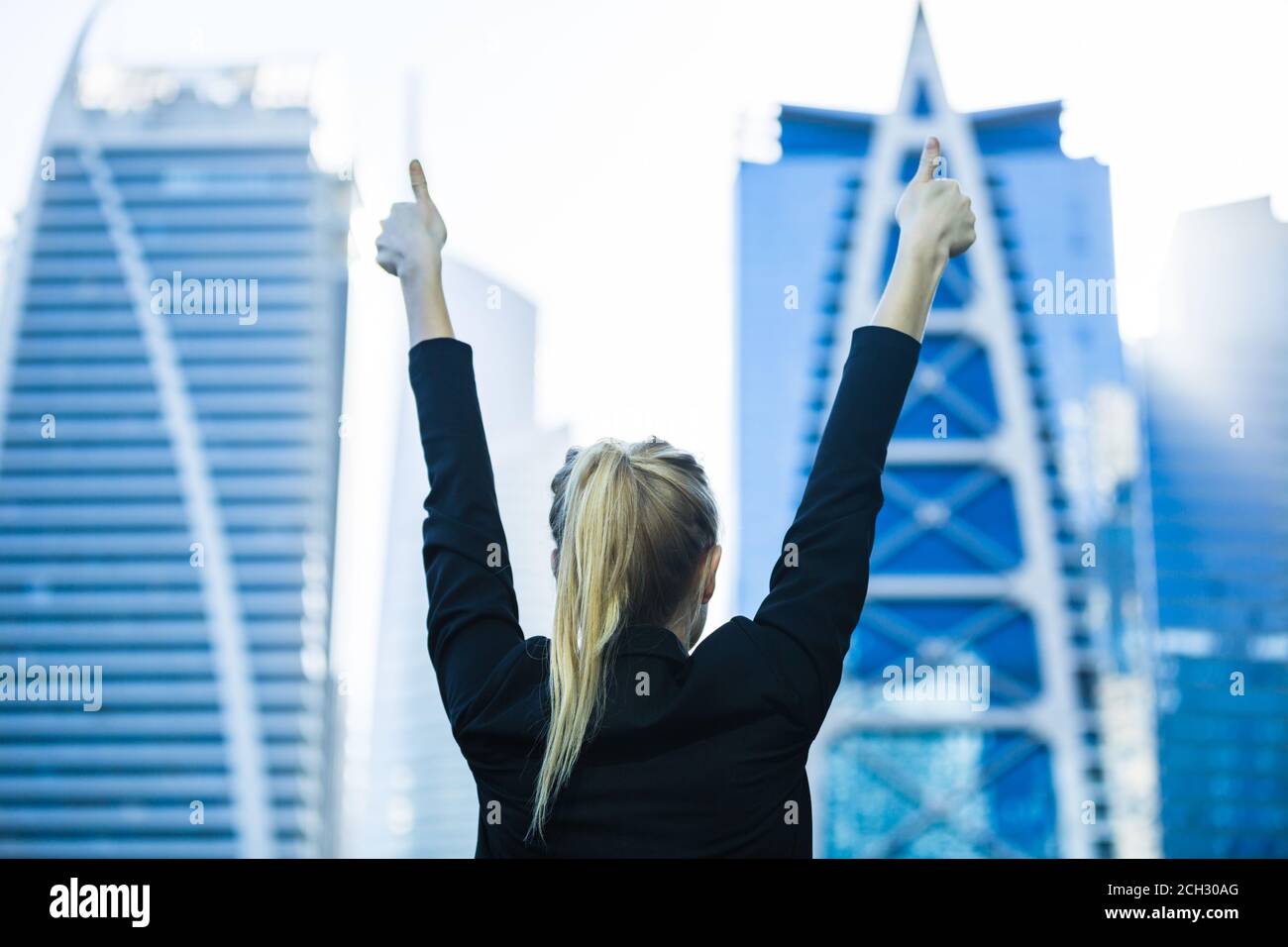 Business success - Celebrating businesswoman overlooking the city high-rise buildings. Optimistic happy woman with hands in the air and thumbs up. Stock Photo