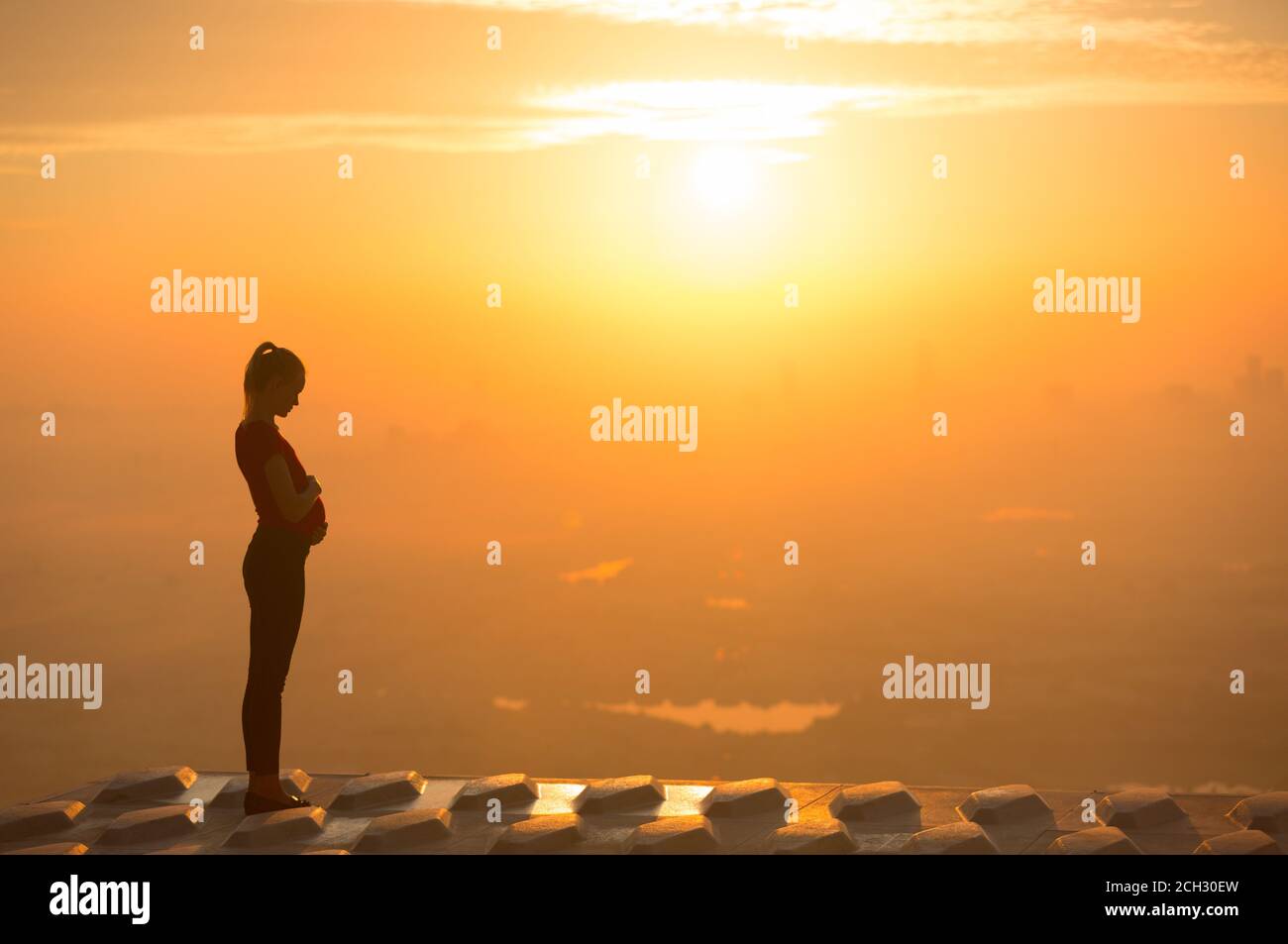 Silhouette of a pregnant woman holding her stomach while standing on a building rooftop during a beautiful orange sunset sky. Maternity and health. Stock Photo