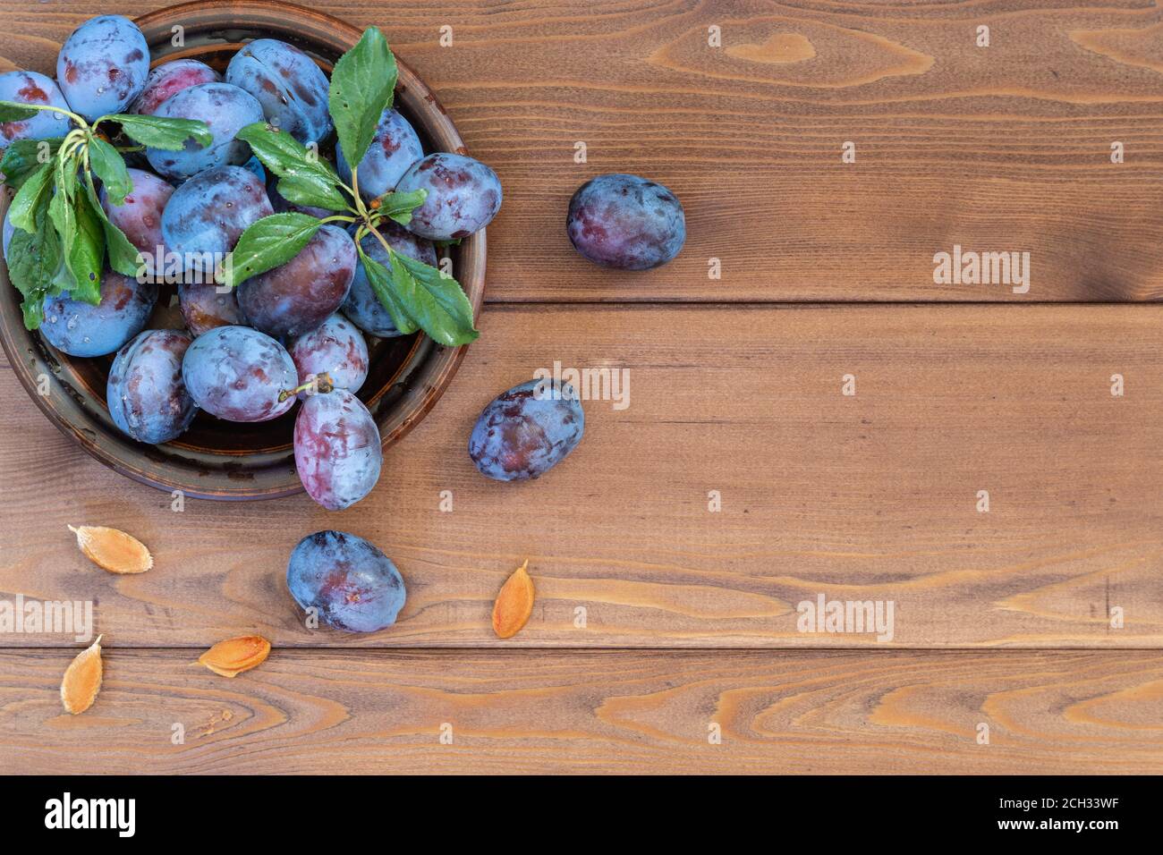 A few washed ripe blue plums with leaves in a brown plate on the wooden table. Several bones lie separately. Copy space. The view from the top Stock Photo