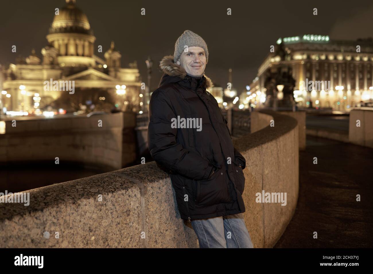 Caucasian man in winter jacket leans on the fence of embankment of Moika river against St. Isaac cathedral in Saint-Petersburg, Russia, and looking to Stock Photo