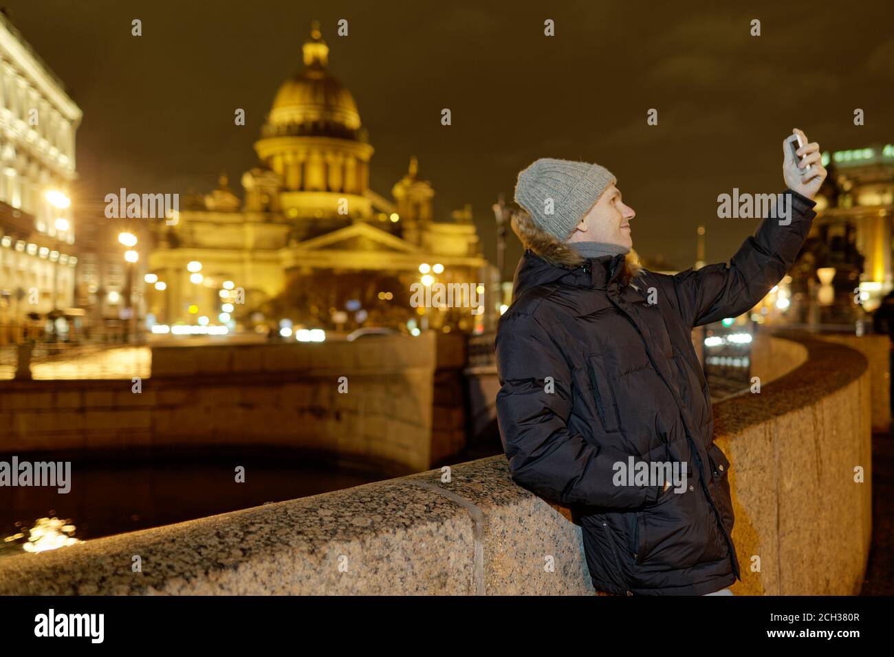Caucasian man in winter jacket makes selfie using his smartphone at the embankment of Moika river against St. Isaac cathedral in Saint-Petersburg, Rus Stock Photo