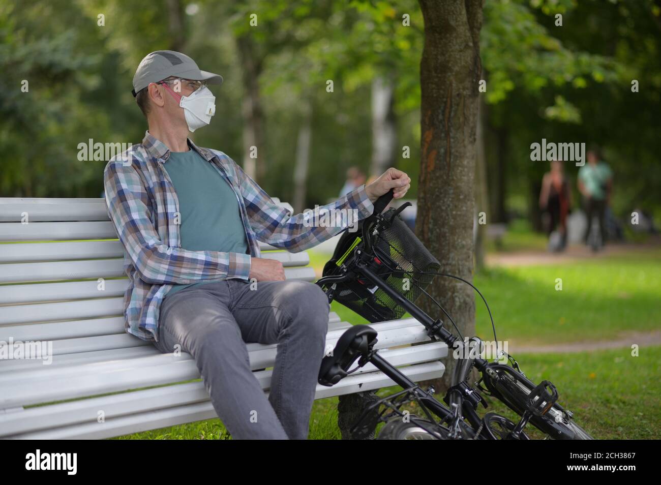 Mature Caucasian man in a respirator with his bicycle resting on a bench in a city park Stock Photo