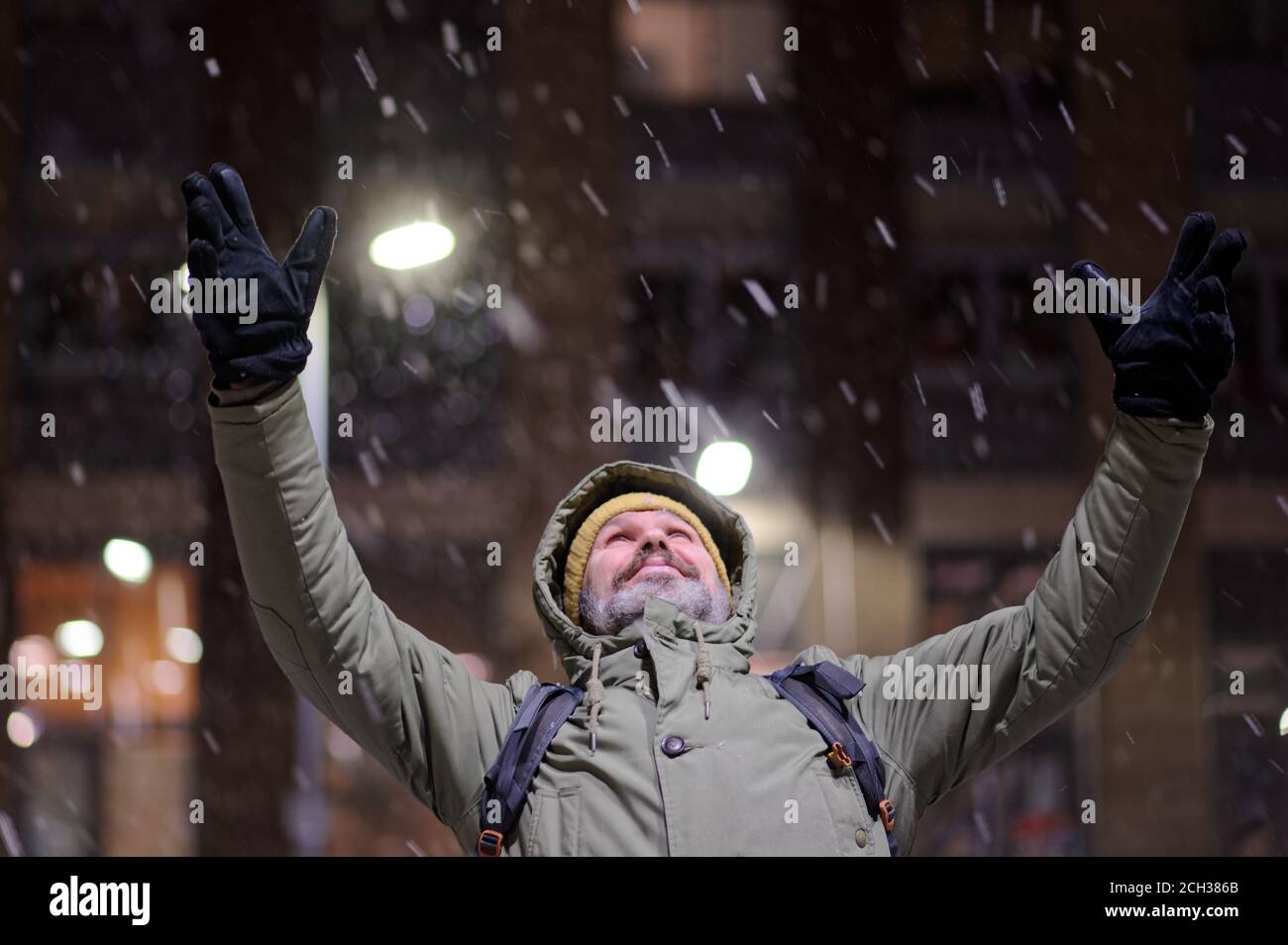 Mature Caucasian bearded man in winter jacket standing under snowfall with hands raised to the sky and looking upward Stock Photo