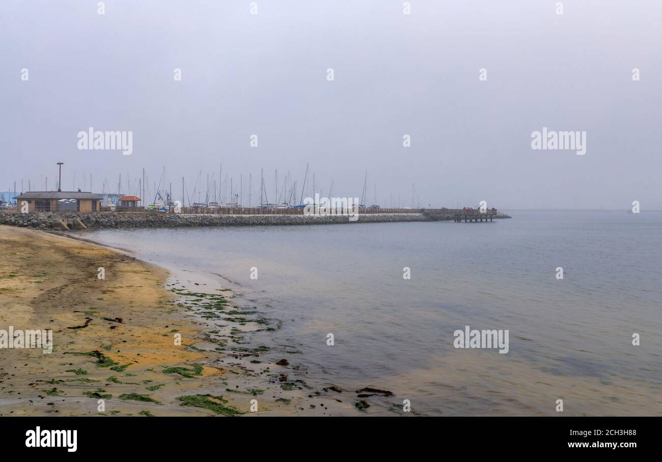 View of a sandy Pacific Ocean beach and a fishing pier in Pillar Point Half Moon Bay near San Francisco on a foggy day, with boats in the background Stock Photo