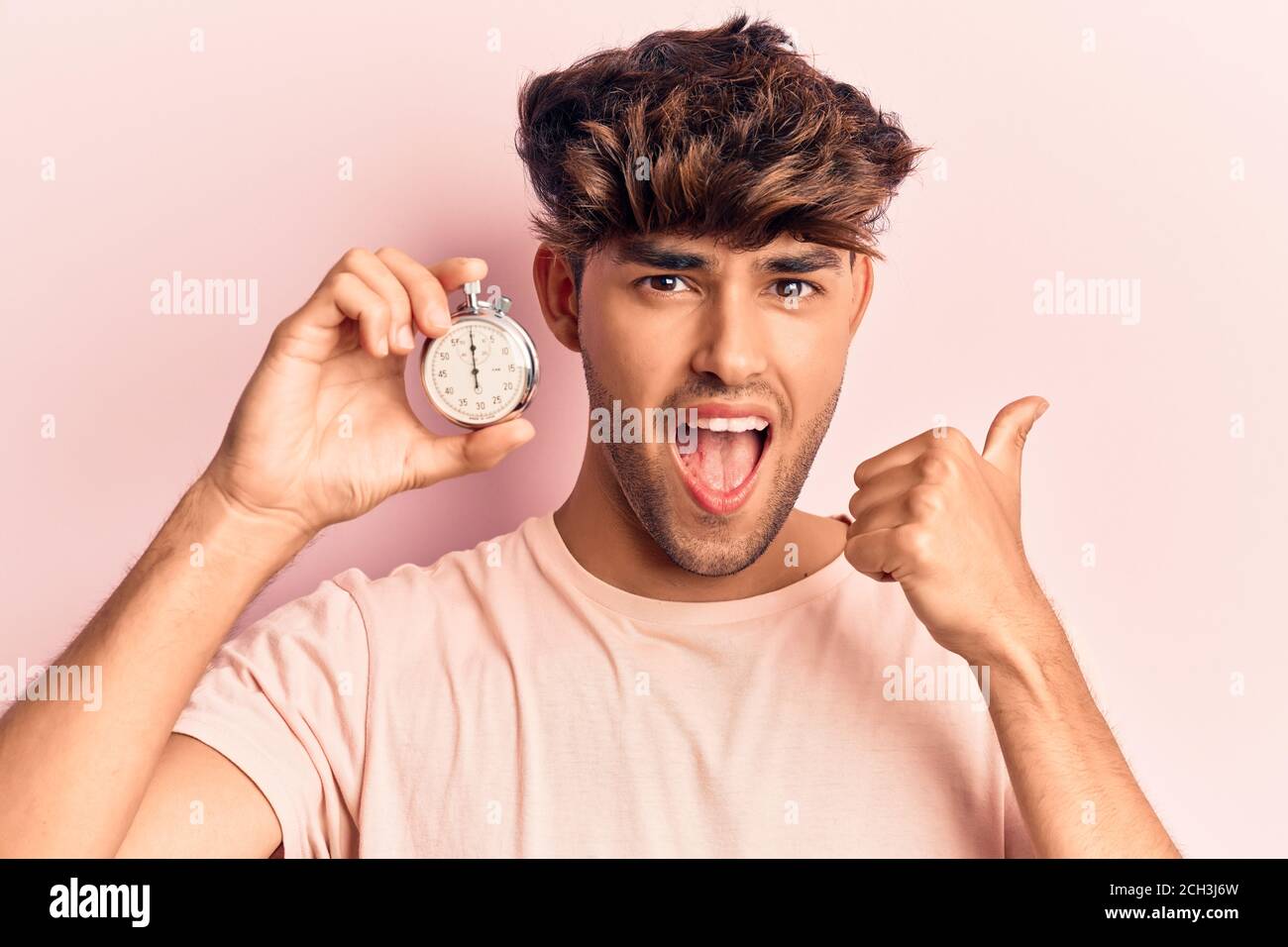 Young hispanic man holding stopwatch pointing thumb up to the side ...