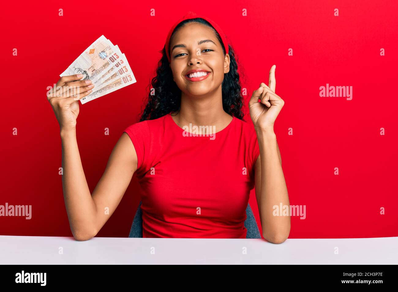 Young african american girl holding united kingdom pounds sitting on the table smiling happy pointing with hand and finger to the side Stock Photo
