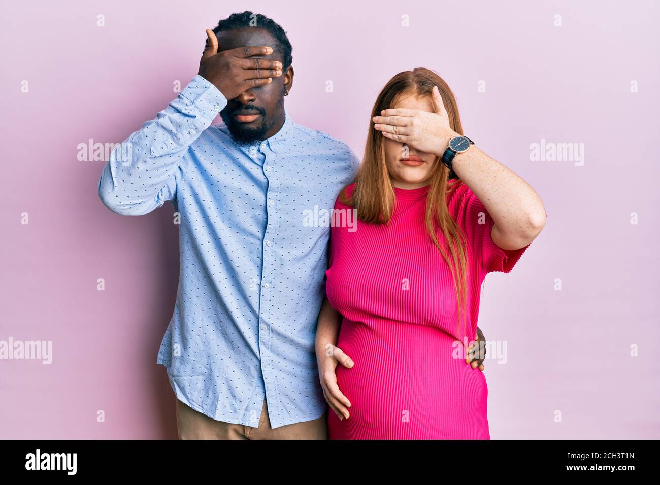 Young interracial couple expecting a baby, touching pregnant belly covering eyes with hand, looking serious and sad. sightless, hiding and rejection c Stock Photo