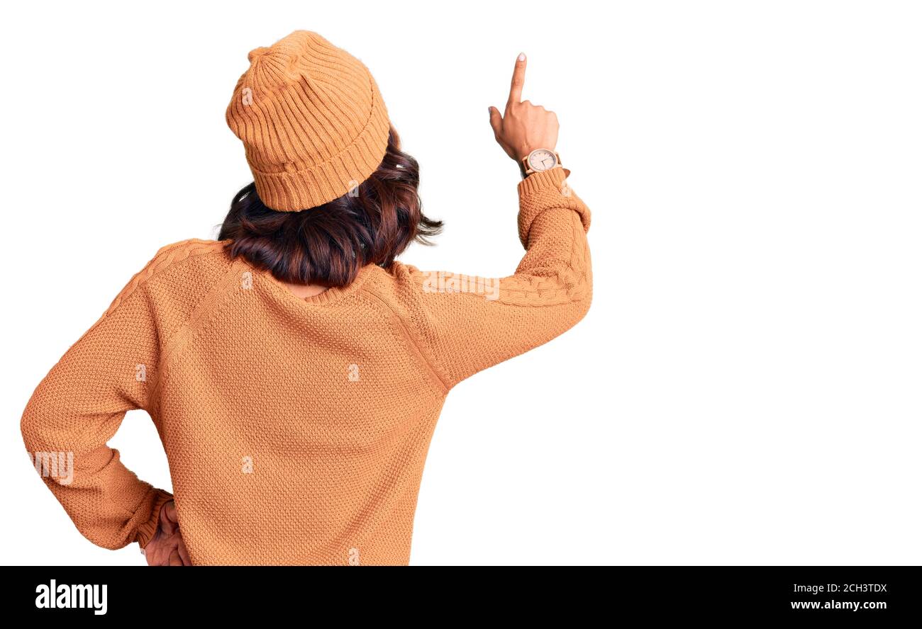 Young beautiful mixed race woman wearing wool sweater and winter hat posing backwards pointing ahead with finger hand Stock Photo