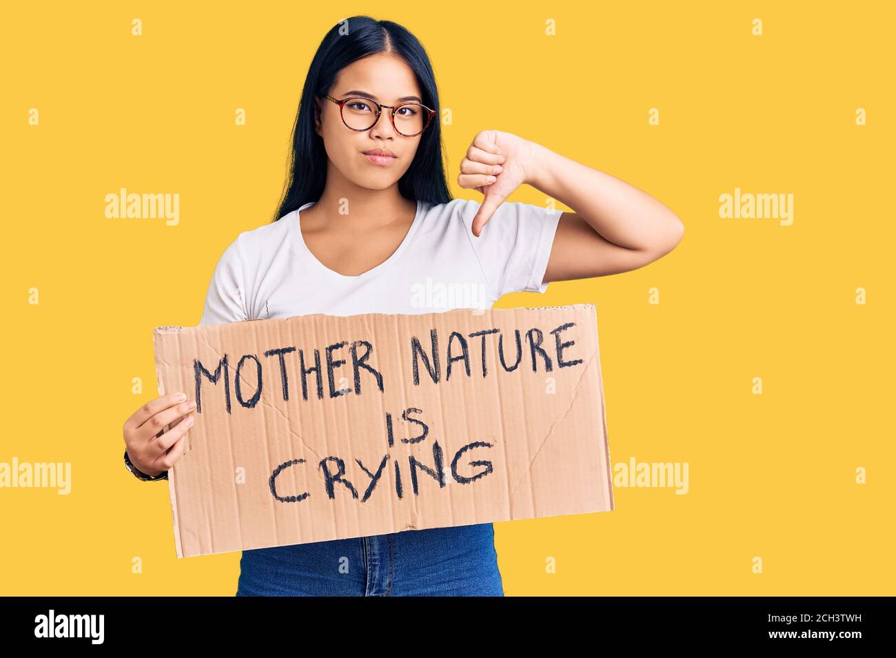 Young beautiful asian girl holding mother nature is crying protest cardboard banner with angry face, negative sign showing dislike with thumbs down, r Stock Photo