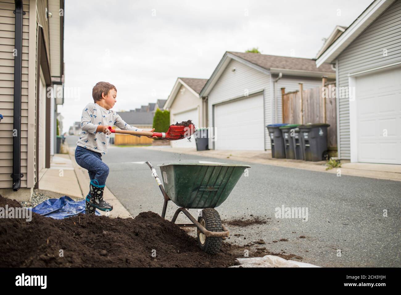 Young boy lifting shovel of dirt into wheelbarrow in back alley Stock Photo