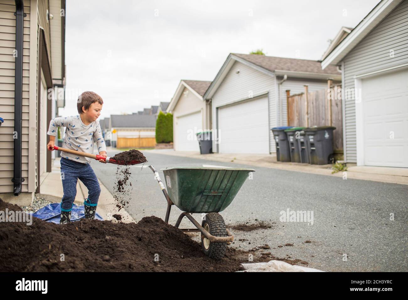 Young boy lifting shovel full of soil into wheelbarrow in back alley Stock Photo