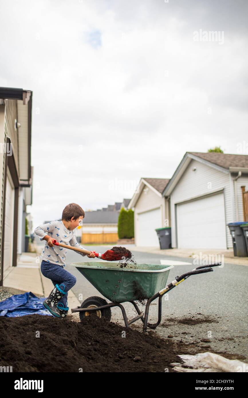 Young boy lifting shovel full of soil into wheelbarrow in back alley ...