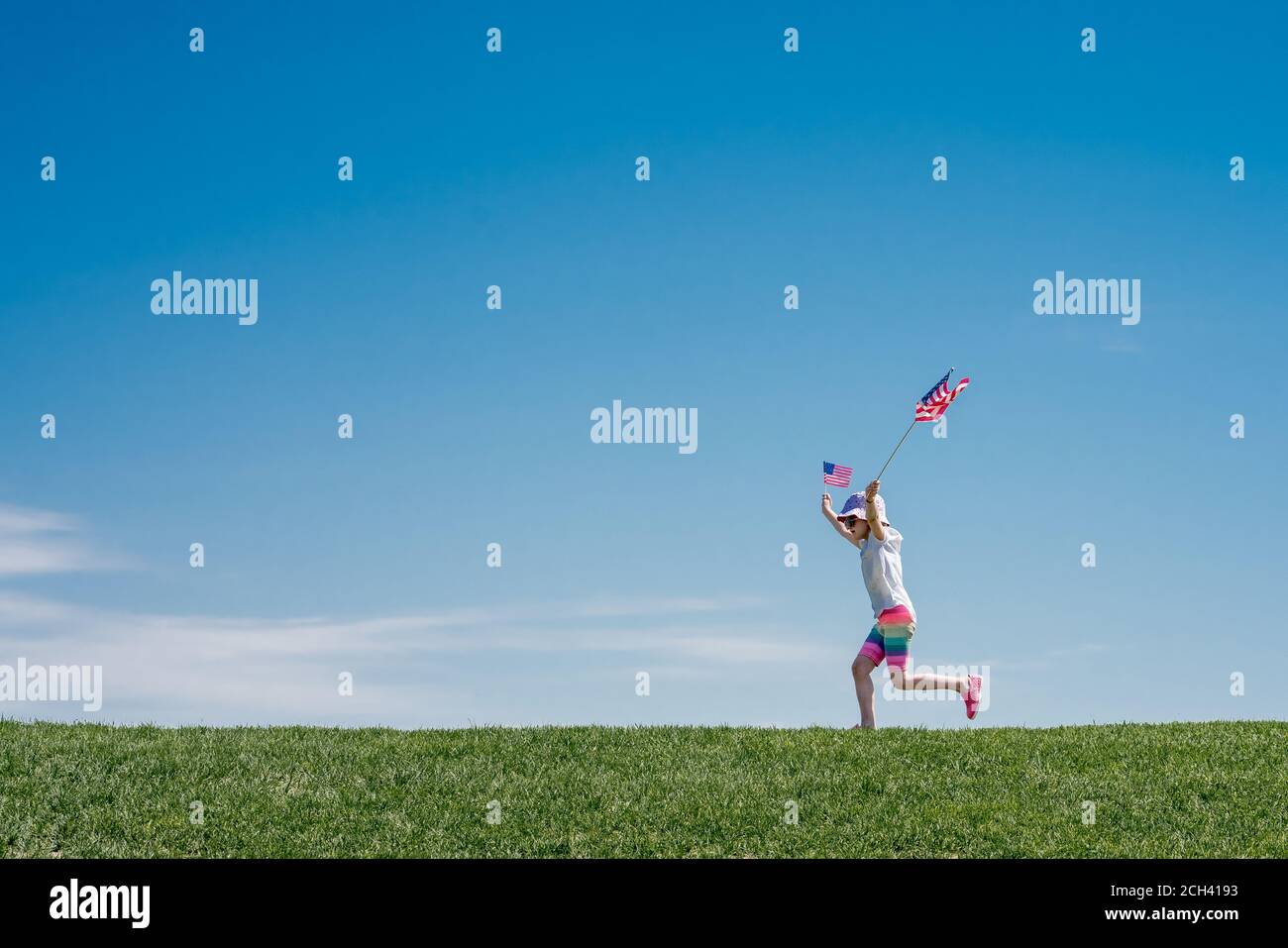 young girl leaps across a grass hill waving 2 American flags Stock Photo
